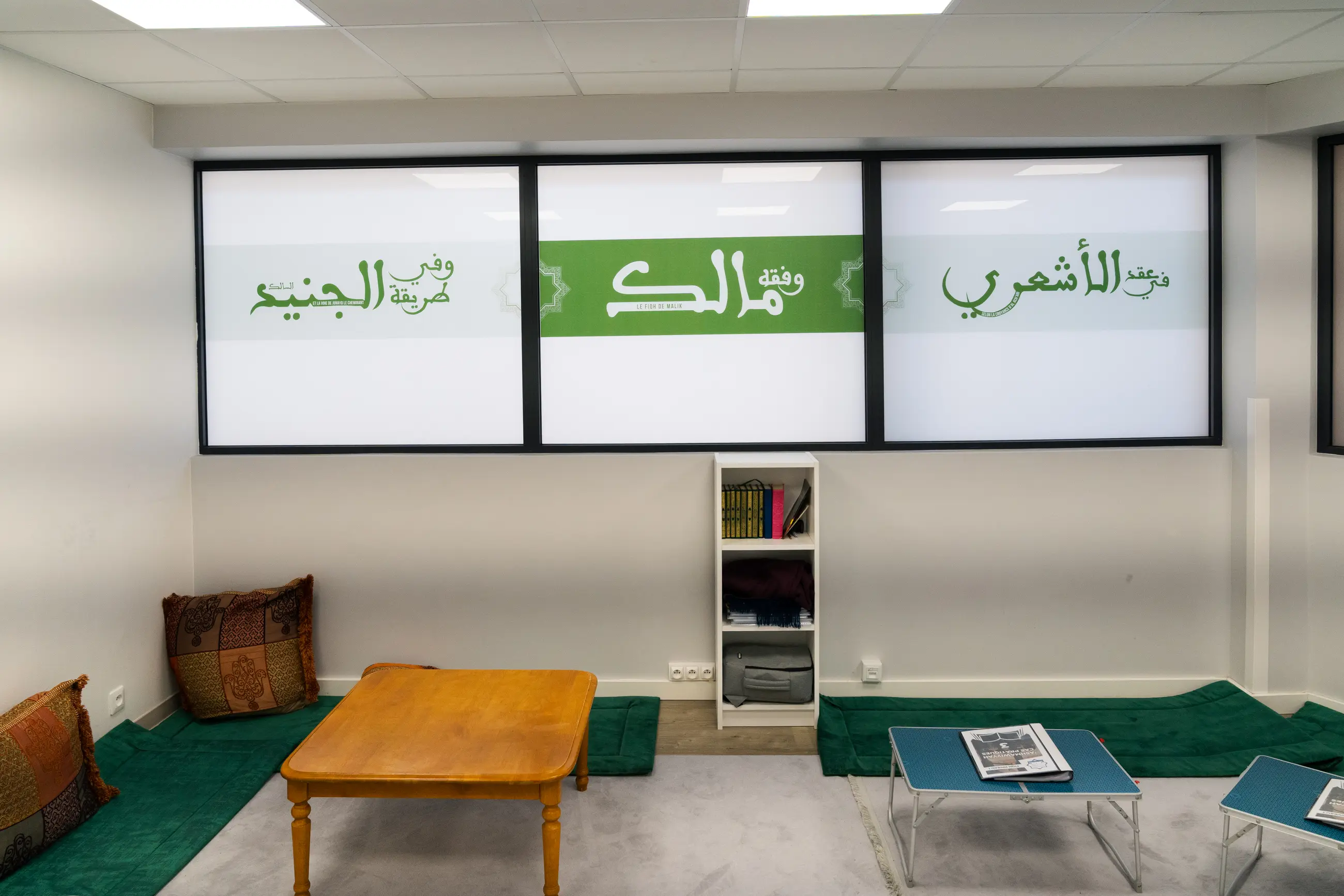 La salle Ibn ʿĀshir de face : la verrière calligraphiée sur toute la largeur, les tapis verts et les tables basses au sol, une étagère de corans sous les fenêtres.