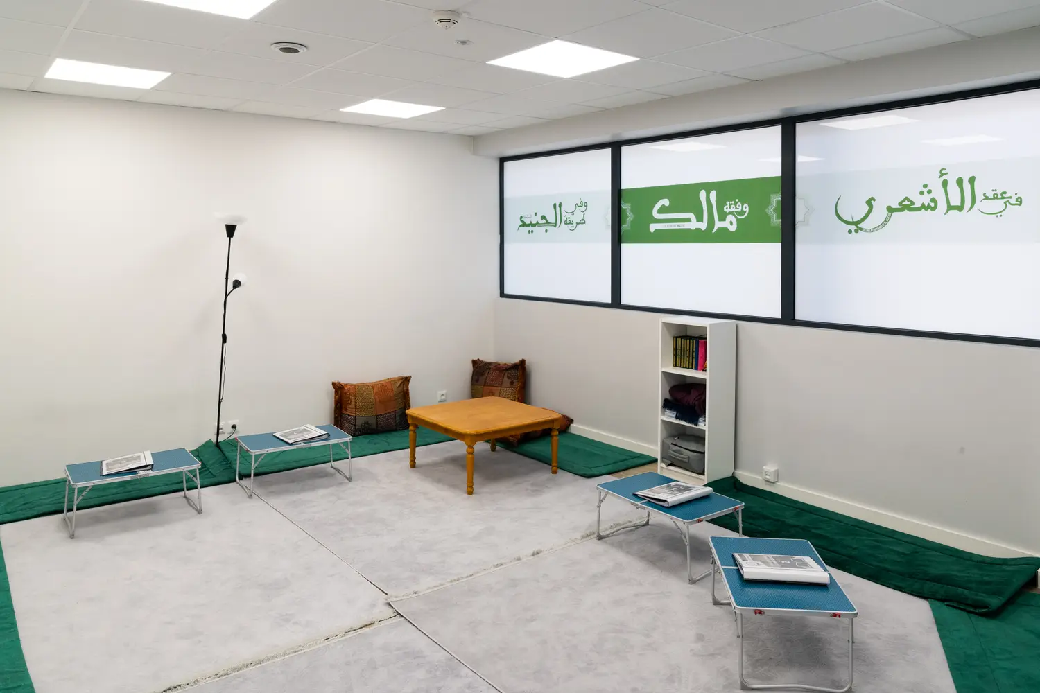 La salle Ibn ʿĀshir vue de l’autre bout : les tapis verts le long des murs, la table basse de l’enseignant et ses deux coussins brodés, l’étagère de corans, et les tables pliantes des étudiants.
