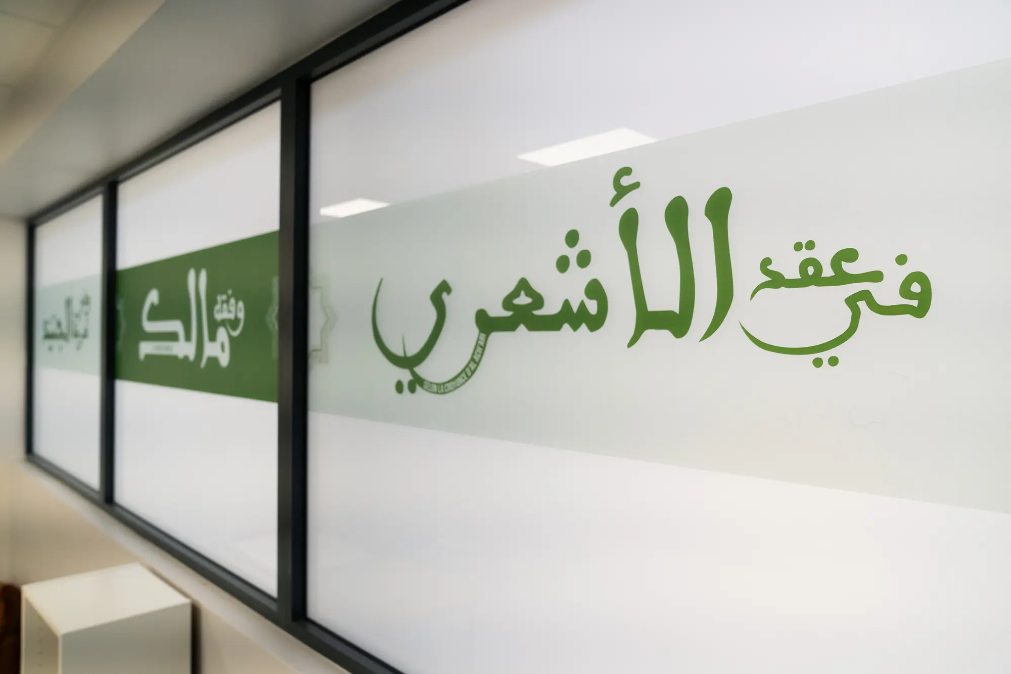 La verrière vue en enfilade : « selon la croyance d’al-Ashʿarī » net au premier plan, « le fiqh de Mālik » qui s’éloigne, le troisième panneau hors champ.