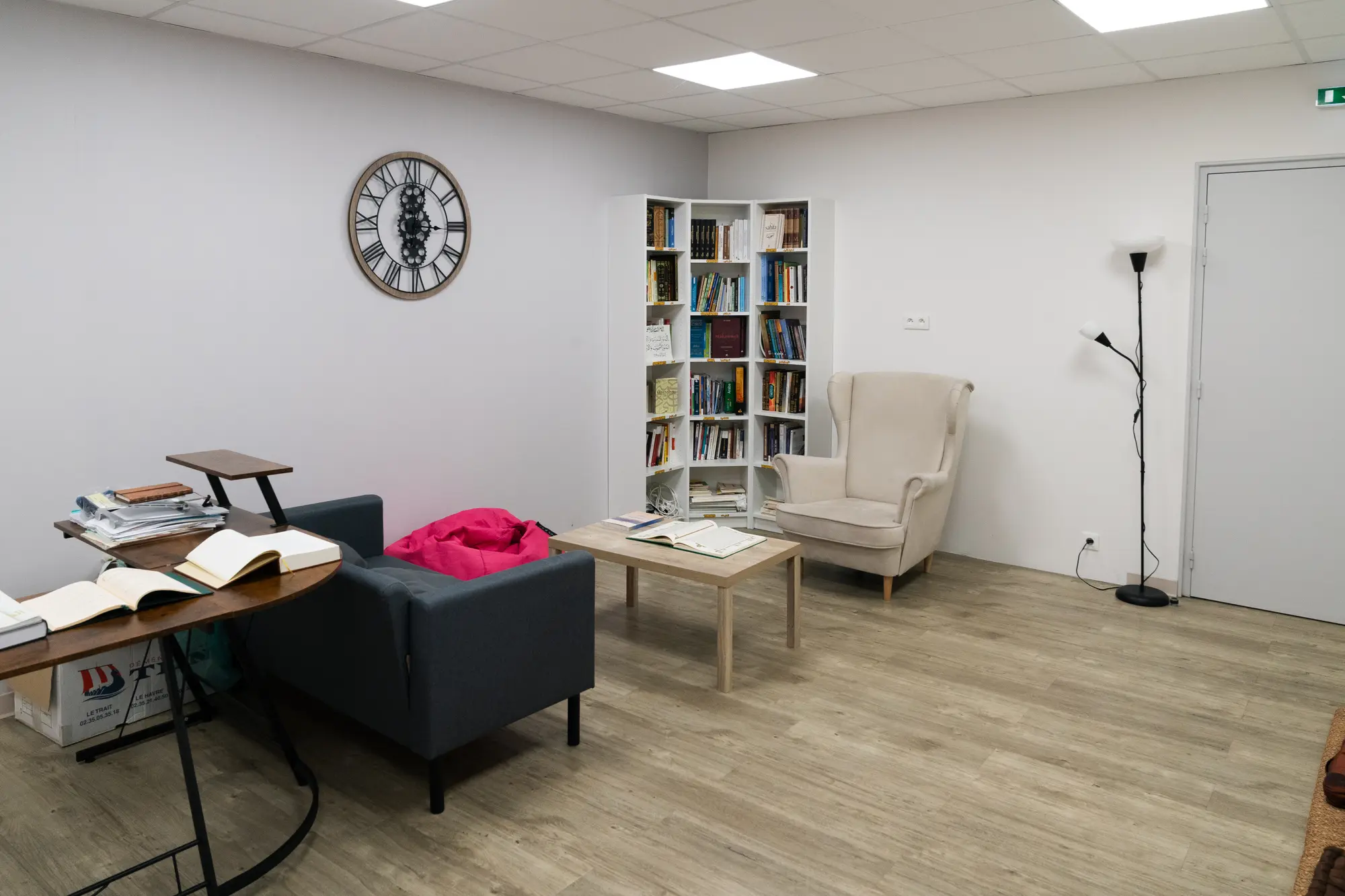 Le coin bibliothèque de la salle Ummunā ʿĀïcha : les rayonnages d’angle, le fauteuil crème, la table basse et son livre ouvert.