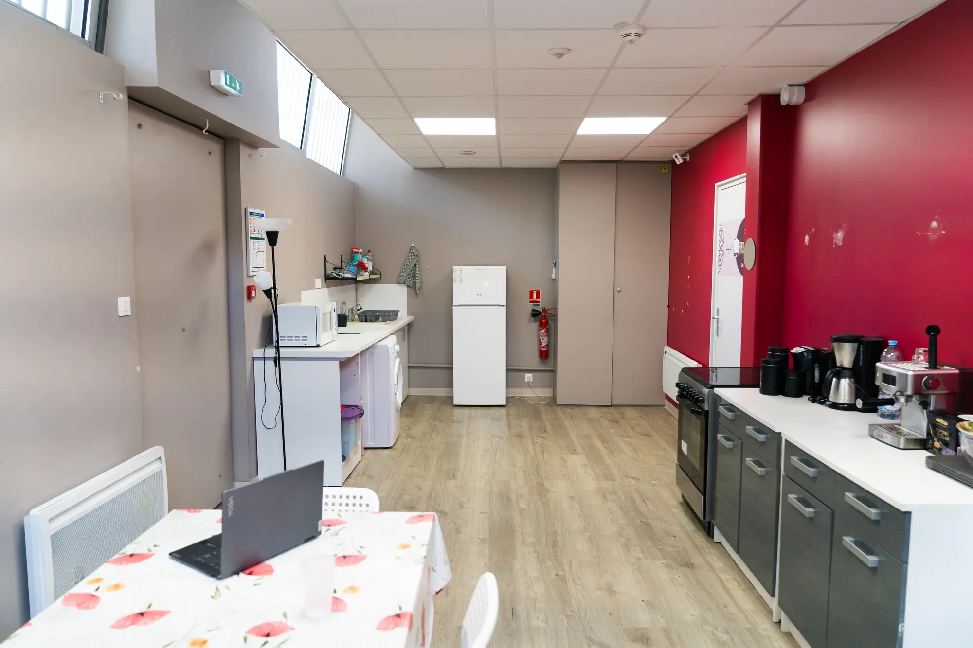 La salle Alhambra depuis l’autre bout : le mur cramoisi et le coin cuisine à droite, les tables à nappe fleurie au centre, un ordinateur portable resté ouvert.