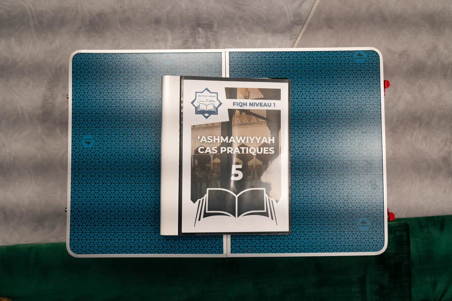 Le fascicule de cas pratiques de la ʿAshmāwiyya, fiqh niveau 1, posé ouvert sur le tapis vert : couverture turquoise au sceau de l’Institut.