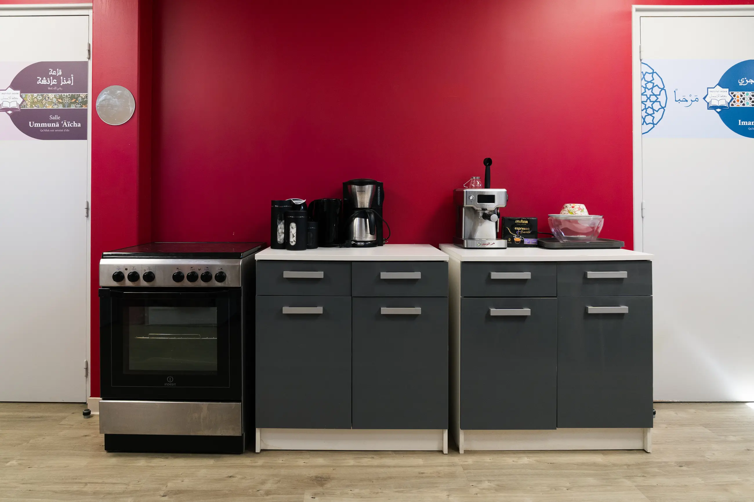 Le coin cuisine, frontal : le mur cramoisi, la cuisinière, la cafetière et la machine à expresso sur le plan de travail, et de part et d’autre les deux portes signées des salles Ummunā ʿĀïcha et Ibn Juzay.