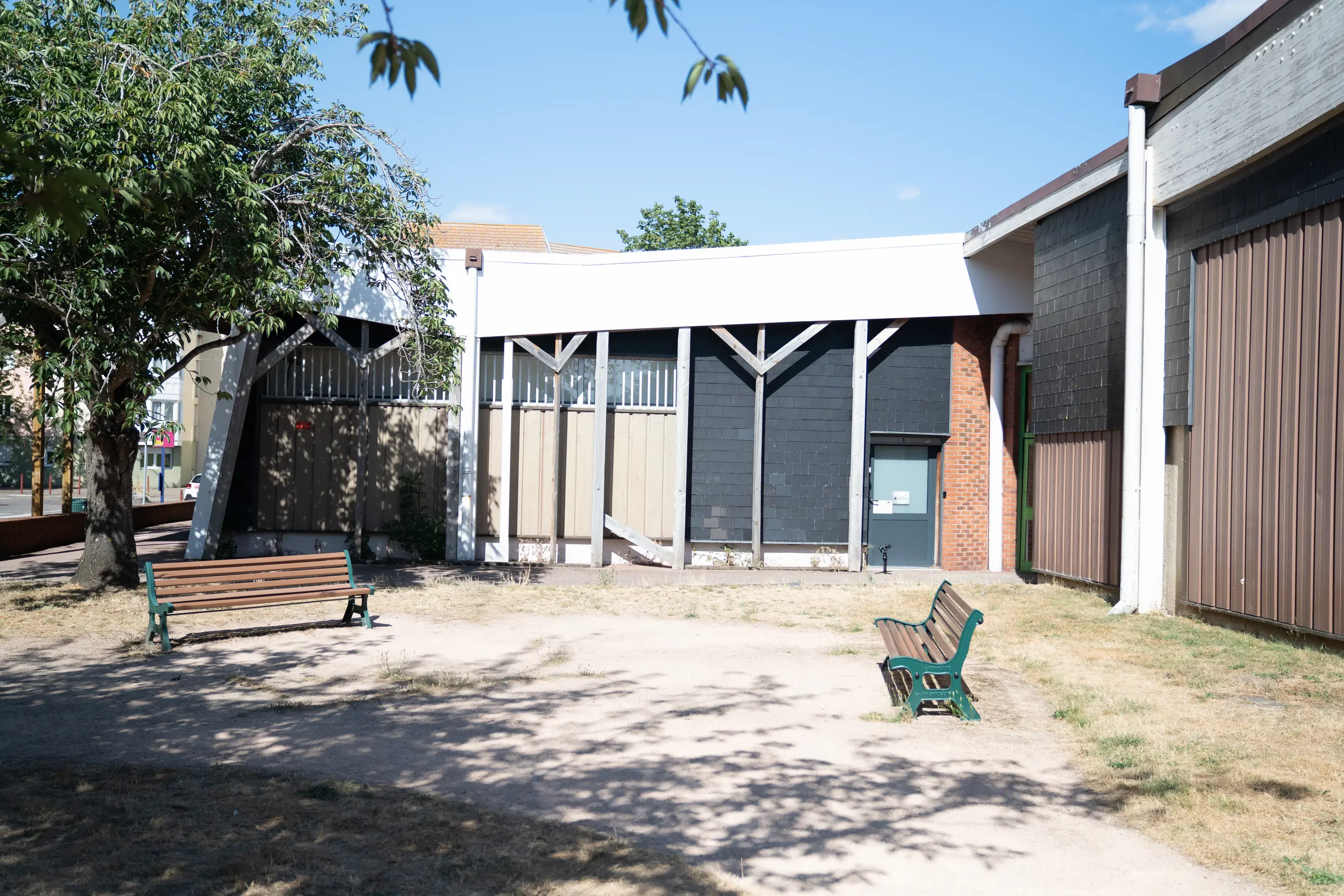 L’extérieur : la cour plantée d’un arbre, deux bancs, et le préau devant les bâtiments de brique du centre commercial des Bruyères.