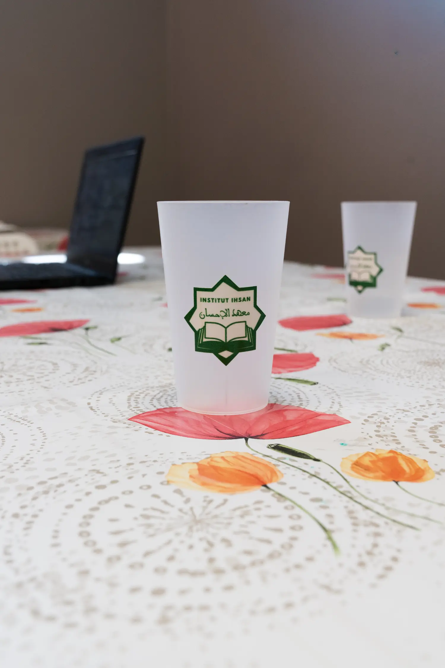 Un gobelet au sceau de l’Institut sur la nappe fleurie, un second flou derrière, et un ordinateur portable resté ouvert.