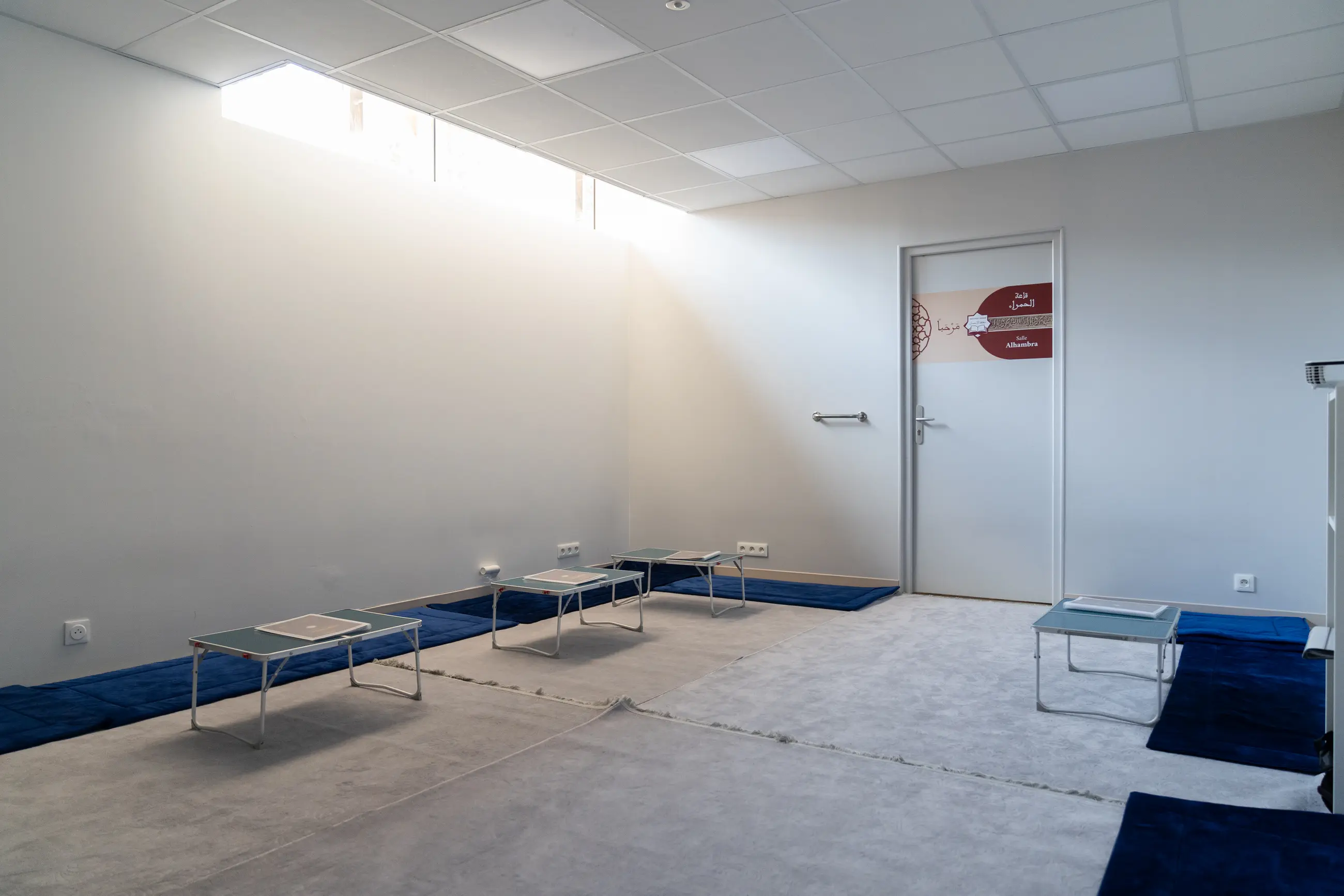 La salle Ibn Juzay : la lumière descend en biais du bandeau de fenêtres, les tapis bleus et les tables basses courent le long des murs, la porte du fond mène à la salle Alhambra.