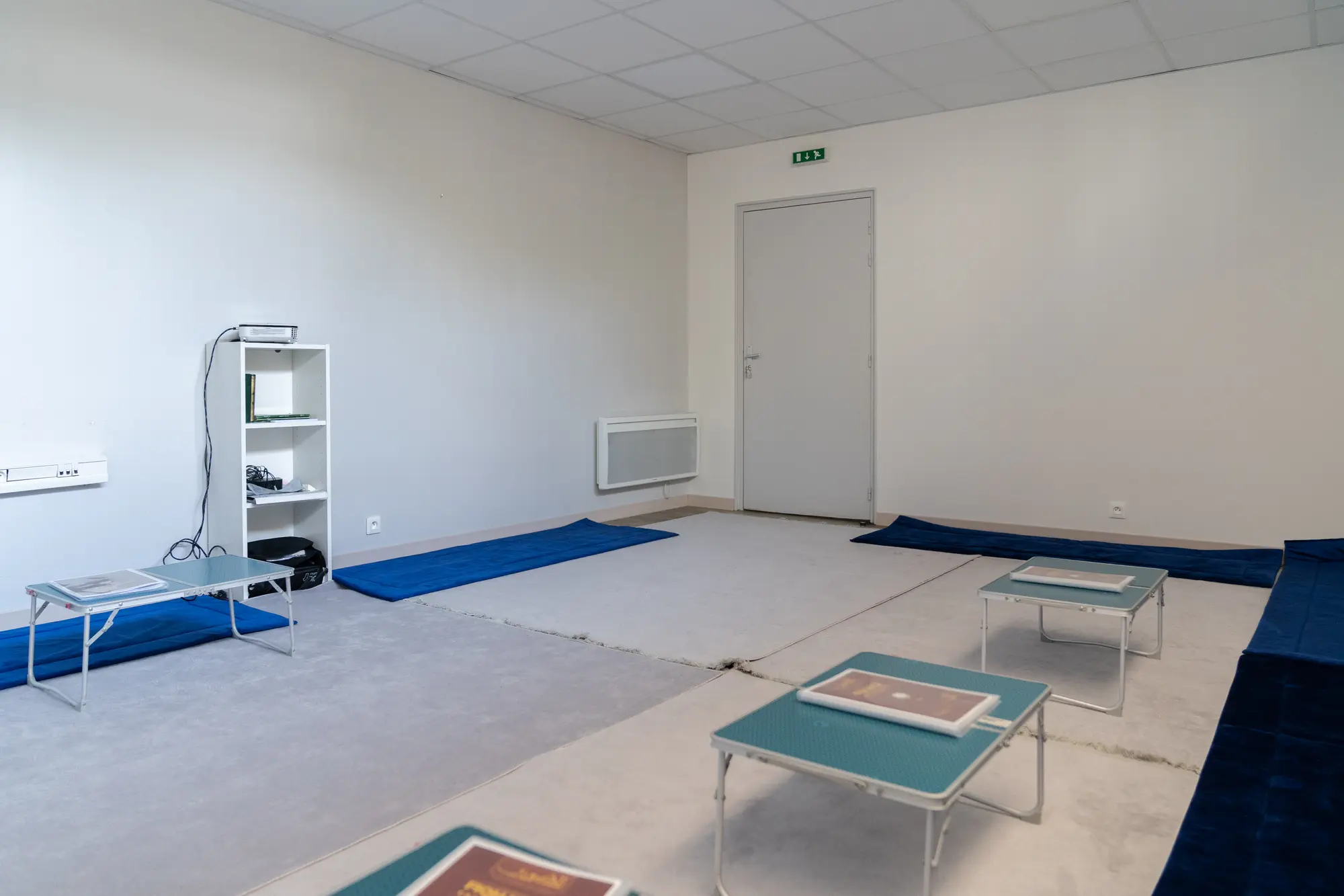 La salle Ibn Juzay de face, vide : les tapis bleus le long des murs blancs, les tables basses à plateau turquoise, et la desserte de corans près de la porte.